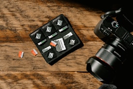 A wooden surface with a camera and a storage pouch containing memory cards. Several SD cards are arranged neatly in the pouch, with a few placed on the table. The camera is positioned to the right, showing part of its lens and control dials.