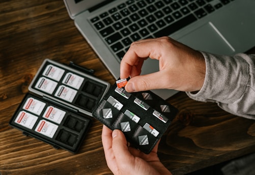 Two hands are holding a black SD card holder filled with multiple SD cards, with a laptop and another open SD card holder on a wooden table.