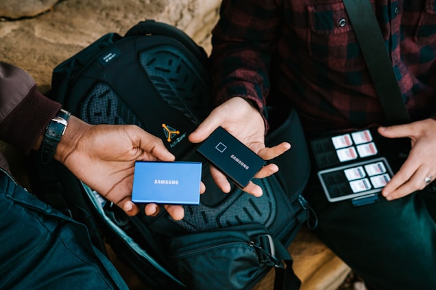 Two individuals exchange portable Samsung SSDs while sitting with a black backpack. One person holds a blue SSD, and the other holds a black SSD. A collection of SD cards is visible next to the person on the right, who is wearing a plaid shirt.