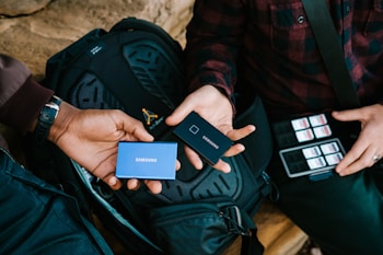 Two individuals exchange portable Samsung SSDs while sitting with a black backpack. One person holds a blue SSD, and the other holds a black SSD. A collection of SD cards is visible next to the person on the right, who is wearing a plaid shirt.