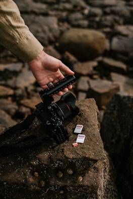A hand adjusting the handle of a professional camera with multiple SD cards placed on a textured rock surface in a natural outdoor setting.