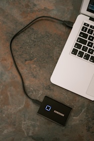 A laptop is connected to a Samsung external solid-state drive via a USB cable. The laptop's keyboard is partially visible on a textured surface.