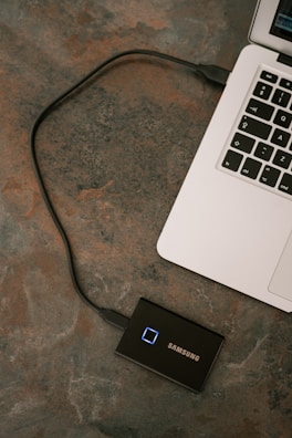 A laptop is connected to a Samsung external solid-state drive via a USB cable. The laptop's keyboard is partially visible on a textured surface.