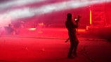 A person holding a professional camera stands on a dimly lit street or outdoor area, which is illuminated with intense red lighting. Fog or smoke effects add a surreal atmosphere. In the background, there's a group of people and a structure with scaffolding, suggesting a concert or event setting.
