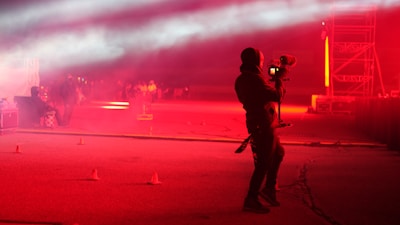 A person holding a professional camera stands on a dimly lit street or outdoor area, which is illuminated with intense red lighting. Fog or smoke effects add a surreal atmosphere. In the background, there's a group of people and a structure with scaffolding, suggesting a concert or event setting.