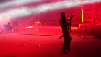 A person holding a professional camera stands on a dimly lit street or outdoor area, which is illuminated with intense red lighting. Fog or smoke effects add a surreal atmosphere. In the background, there's a group of people and a structure with scaffolding, suggesting a concert or event setting.