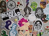 A collage of stickers featuring various graphic designs, including skulls, skeletons, a green hand with an eyeball, a dancing figure with a rose head, and other random whimsical and gothic artwork. One sticker prominently reads 'Planet of the Dead'.