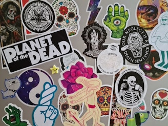 A collage of stickers featuring various graphic designs, including skulls, skeletons, a green hand with an eyeball, a dancing figure with a rose head, and other random whimsical and gothic artwork. One sticker prominently reads 'Planet of the Dead'.