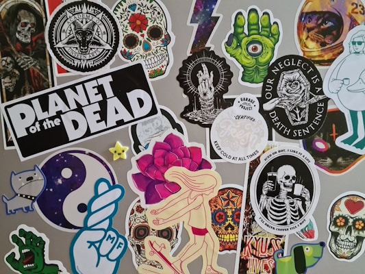 A collage of stickers featuring various graphic designs, including skulls, skeletons, a green hand with an eyeball, a dancing figure with a rose head, and other random whimsical and gothic artwork. One sticker prominently reads 'Planet of the Dead'.