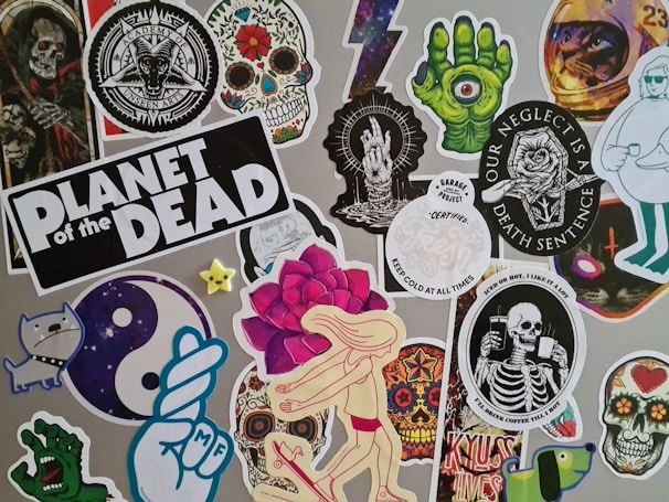 A collage of stickers featuring various graphic designs, including skulls, skeletons, a green hand with an eyeball, a dancing figure with a rose head, and other random whimsical and gothic artwork. One sticker prominently reads 'Planet of the Dead'.