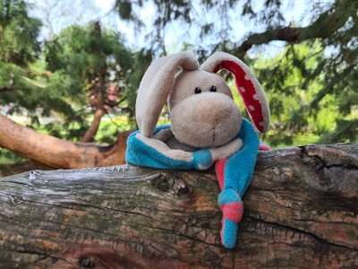 A plush toy rabbit with long ears sits on a textured wooden surface. The rabbit has a soft, beige face with black eyes and a pink nose. Its ears are lined with red fabric featuring white polka dots. The rabbit's body is covered in blue and pink fabric, giving it a colorful appearance. The background consists of lush green trees with sunlight filtering through, creating a serene outdoor setting.