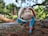 A plush toy rabbit with long ears sits on a textured wooden surface. The rabbit has a soft, beige face with black eyes and a pink nose. Its ears are lined with red fabric featuring white polka dots. The rabbit's body is covered in blue and pink fabric, giving it a colorful appearance. The background consists of lush green trees with sunlight filtering through, creating a serene outdoor setting.