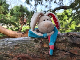 A plush toy rabbit with long ears sits on a textured wooden surface. The rabbit has a soft, beige face with black eyes and a pink nose. Its ears are lined with red fabric featuring white polka dots. The rabbit's body is covered in blue and pink fabric, giving it a colorful appearance. The background consists of lush green trees with sunlight filtering through, creating a serene outdoor setting.