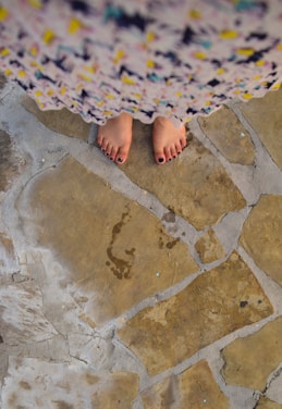 A pair of bare feet with dark-painted toenails stands on a patterned stone floor. The stones vary in shades of beige and brown, with visible grout lines. The feet are partially covered by a colorful, patterned garment, which appears slightly blurred in the foreground.
