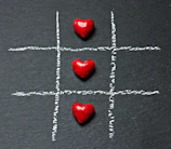 Three red heart-shaped objects are placed in a row on a chalk-drawn tic-tac-toe board on a dark surface.