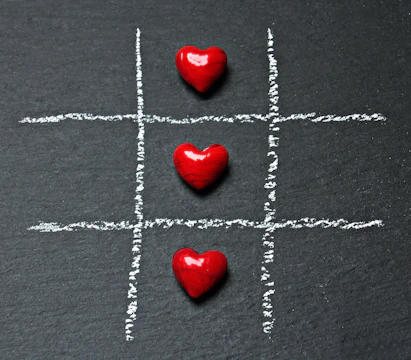 Three red heart-shaped objects are placed in a row on a chalk-drawn tic-tac-toe board on a dark surface.
