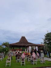 A traditional outdoor ceremony taking place in front of a structure with a brown tiled roof, perhaps a pavilion. Guests are seated on white chairs on a well-manicured lawn, wearing a mix of traditional and formal attire. The setting appears peaceful, surrounded by greenery and under a cloudy sky.