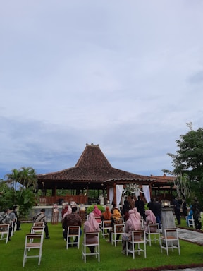 A traditional outdoor ceremony taking place in front of a structure with a brown tiled roof, perhaps a pavilion. Guests are seated on white chairs on a well-manicured lawn, wearing a mix of traditional and formal attire. The setting appears peaceful, surrounded by greenery and under a cloudy sky.