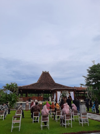 A traditional outdoor ceremony taking place in front of a structure with a brown tiled roof, perhaps a pavilion. Guests are seated on white chairs on a well-manicured lawn, wearing a mix of traditional and formal attire. The setting appears peaceful, surrounded by greenery and under a cloudy sky.