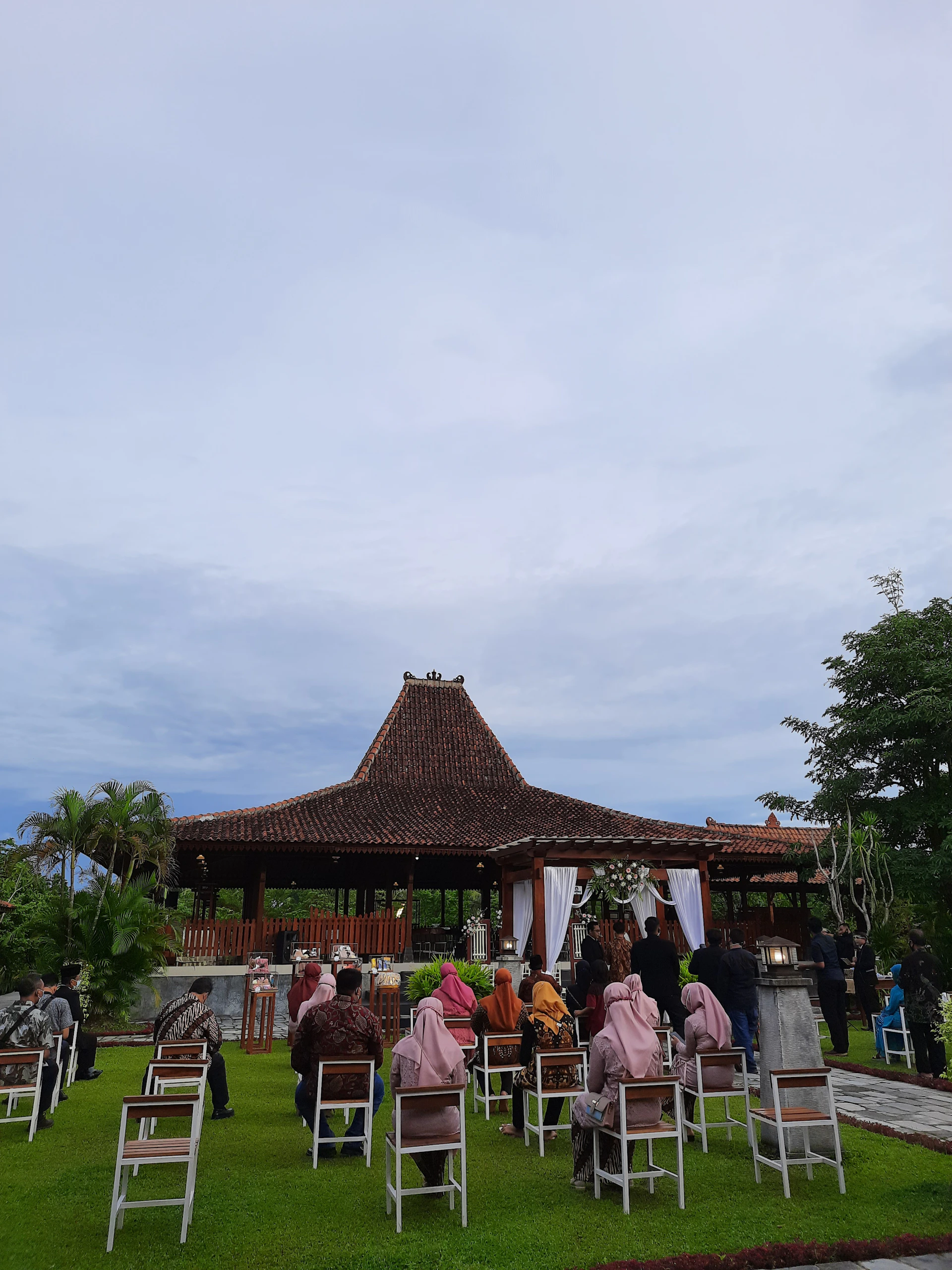 A wide shot of a sunlit outdoor wedding ceremony with guests seated, framed by delicate floral arrangements.