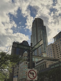 A cityscape with tall buildings reaching into a partly cloudy sky. A street sign marks the intersection of E Chicago Ave and N Michigan Ave, with surrounding architecture showcasing modern and historic styles. Lush green trees are visible in the foreground.