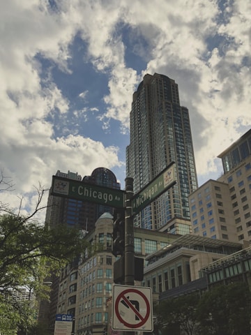 A cityscape with tall buildings reaching into a partly cloudy sky. A street sign marks the intersection of E Chicago Ave and N Michigan Ave, with surrounding architecture showcasing modern and historic styles. Lush green trees are visible in the foreground.