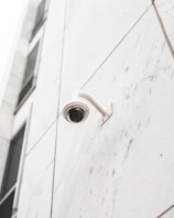 A security camera is mounted on the corner of a modern building with large, light-colored, rectangular panels. The facade has a minimalistic design with clean lines, and parts of window reflections are visible in the upper section.