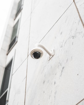 A security camera is mounted on the corner of a modern building with large, light-colored, rectangular panels. The facade has a minimalistic design with clean lines, and parts of window reflections are visible in the upper section.