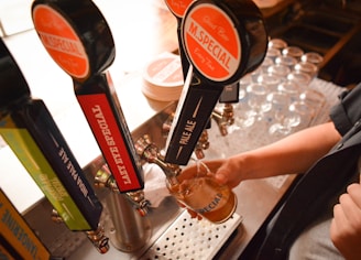 Several beer taps are lined up in a bar setting, with a person pouring a beer into a glass from one labeled 'M. Special'. The counter has a stack of coasters and a tray of empty glasses.