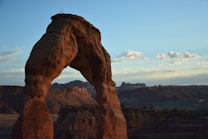 A natural stone arch formation stands prominently in a vast desert landscape. The sky is clear with a few scattered clouds, and the warm evening light casts a soft glow on the rugged terrain.