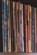 A row of vertically arranged magazines or booklets with varying colorful covers and visible spines lined up side by side. The collection exhibits a mixture of patterns and designs, with some covers worn and slightly bent, indicating frequent handling.