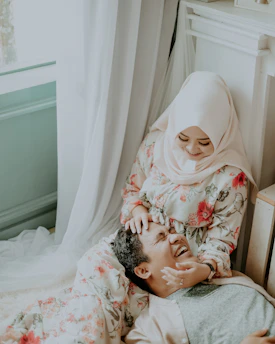 A woman wearing a floral-patterned dress and a light-colored hijab is sitting on the floor next to a man who is lying down with his head on her lap. Both appear to be laughing and sharing a joyful moment together. The room is softly lit, with curtains in the background and a window partially visible.