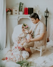A couple is in a cozy, well-decorated room. One person is sitting on a white chair while the other sits on the floor. There are flowers, a table with decor items like potted plants, a small red phone booth, and bird cages. The couple is smiling at each other, holding hands. The room has a soft, romantic ambiance with sheer white curtains and a light color scheme.