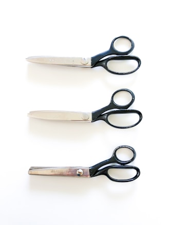 Three pairs of scissors are lined up vertically against a white background. Each pair has shiny metallic blades and black handles. The scissors display subtle differences in blade shape and handle style.