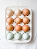 A white ceramic egg tray holding twelve eggs of varying colors, including brown, beige, and light blue. The tray is placed on a white surface.