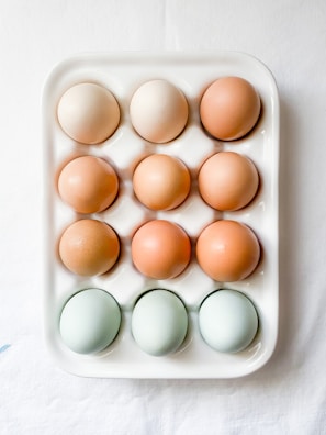 A white ceramic egg tray holding twelve eggs of varying colors, including brown, beige, and light blue. The tray is placed on a white surface.