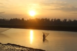 A fisherman holding a large fish by the riverbank during sunset.