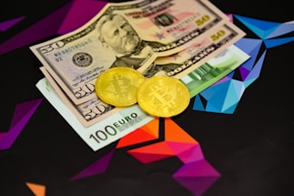 Two physical Bitcoin coins are placed next to a mix of currency bills, including a 100 Euro note and two 50 US Dollar bills. The background features a geometric, colorful pattern with vibrant shades of purple, blue, and orange.