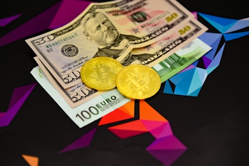 Two physical Bitcoin coins are placed next to a mix of currency bills, including a 100 Euro note and two 50 US Dollar bills. The background features a geometric, colorful pattern with vibrant shades of purple, blue, and orange.
