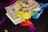 Two physical Bitcoin coins are placed next to a mix of currency bills, including a 100 Euro note and two 50 US Dollar bills. The background features a geometric, colorful pattern with vibrant shades of purple, blue, and orange.