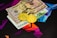 Two physical Bitcoin coins are placed next to a mix of currency bills, including a 100 Euro note and two 50 US Dollar bills. The background features a geometric, colorful pattern with vibrant shades of purple, blue, and orange.