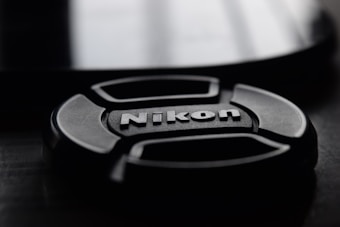 A close-up view of a Nikon camera lens cap placed on a dark surface. The lighting creates a soft reflection on the surface, highlighting the brand's name in the center of the cap.