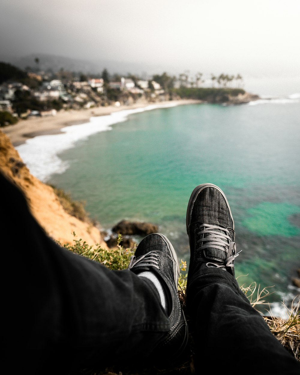 person in black pants and black sneakers sitting on brown rock near body of water during