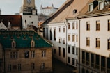 A European cityscape featuring historical architecture with a prominent green-roofed building in the foreground. The scene includes ornate roofs, a clock tower, and a distant castle on a hill. Sunlight creates contrasting shadows on the beige walls of the buildings.