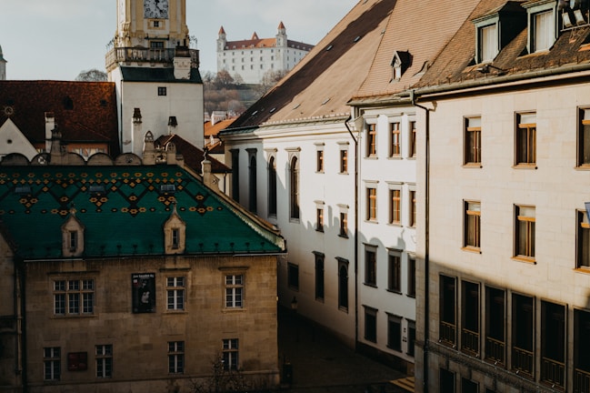 A European cityscape featuring historical architecture with a prominent green-roofed building in the foreground. The scene includes ornate roofs, a clock tower, and a distant castle on a hill. Sunlight creates contrasting shadows on the beige walls of the buildings.