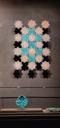 A display of intricately designed tiles with geometric and floral patterns. The tiles are arranged in a symmetrical pattern on a wall, featuring a blend of turquoise and earthy tones. A leaf-shaped ceramic ornament sits on a stand below, matching the tiles' color scheme.