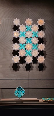 A display of intricately designed tiles with geometric and floral patterns. The tiles are arranged in a symmetrical pattern on a wall, featuring a blend of turquoise and earthy tones. A leaf-shaped ceramic ornament sits on a stand below, matching the tiles' color scheme.