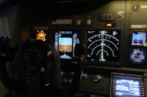 The image features the cockpit of an aircraft, displaying various control panels, illuminated instruments, dials, and screens showing flight data. Visible are the yoke and multiple indicators for navigation and system status, with labels and lights.