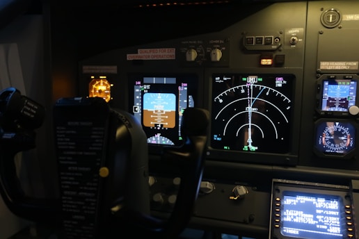 The image features the cockpit of an aircraft, displaying various control panels, illuminated instruments, dials, and screens showing flight data. Visible are the yoke and multiple indicators for navigation and system status, with labels and lights.