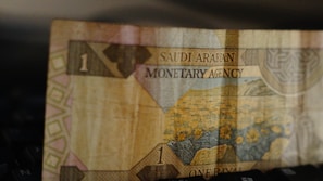 A close-up view of a paper currency note from Saudi Arabia, featuring floral imagery and intricate patterns. The note has the denomination of one rial, with the text 'Saudi Arabian Monetary Agency' printed on it.
